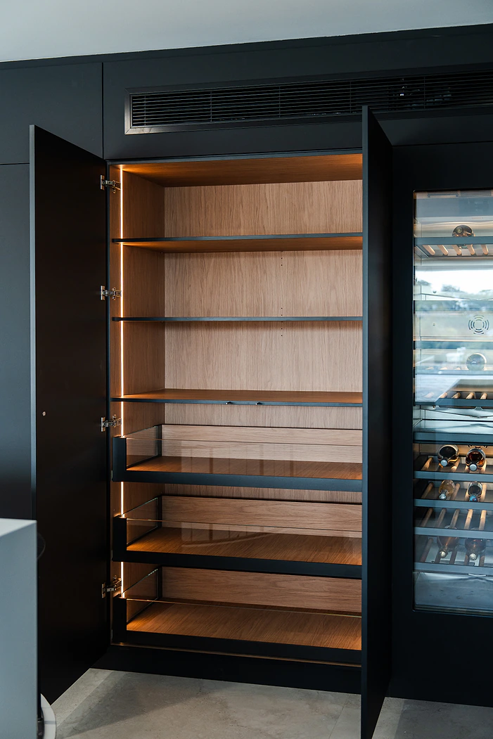 Butler’s pantry with lit timber shelves behind black doors